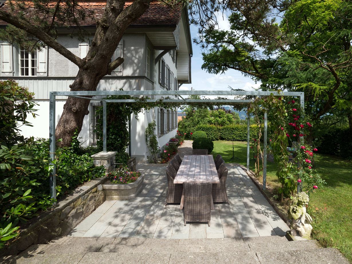 Terrasse Kandla Grey en grès avec un salon de jardin en bois, une fontaine fleurie et de la verdure tout autour